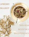 A cup of Raspberry Earl White Traveler tea with tea leaves floating inside is shown from above. Loose tea leaves are scattered on the surface beside the cup. The text reads: "Raspberry Earl White Traveler," "Skin Health," "Boost Mood," and "Brightens Mood." Experience the enchantment of Magic Hour Organic Tea.