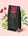 A black tea bag labeled "Raspberry Earl Grey Black Tea" stands upright, surrounded by illustrated raspberry fruits, leaves, and white tea flowers on a soft pink background. Crafted with biodynamically grown Ceylon black tea and infused with organic freeze-dried raspberries, this blend captures the vibrant essence of nature in every cup.