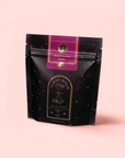 A black and purple resealable pouch labeled "Magic Hour" with gold stars displays "Raspberry Earl White Tea," infused with bergamot and antioxidants, standing upright on a light pink background.