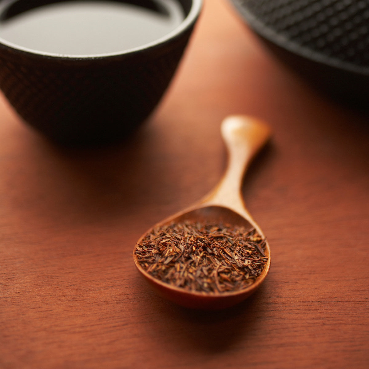 A wooden spoon filled with dried tea leaves is placed on a wooden surface. Behind the spoon, there are two black tea cups, one of which is in focus and appears to be filled with liquid.