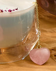 A clear glass cup containing a frothy beverage sits on a wooden surface. The drink is adorned with dried rose petals, and a chain with the Rose Quartz Heart Tea Strainer, featuring a pink heart-shaped pendant, dangles from the cup.