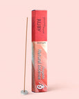A box of Shoyeido Natural Incense : Ruby, renowned for its rich sandalwood aroma, stands upright beside a lit incense stick in a small round holder, set against a pale pink background.