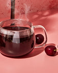 A steaming glass mug brimming with Heart Health Tea Sampler Set sits on a pink surface, accompanied by three fresh cherries. The backdrop showcases a warm, textured pink wall, fostering a cozy ambiance that suggests benefits for heart health and cardiovascular wellness.