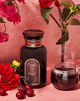 A dark apothecary jar of Best Sellers of Magic Hour Trio Box labeled “Ruby Moon” sits next to a glass mug of steaming dark tea, surrounded by red flowers and cherries.