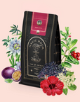 A black and gold tea bag labeled “Ruby Moon™ : Hibiscus Elderberry Tea” is surrounded by illustrated passionfruit, hibiscus, elderberry, and leaves—highlighting this caffeine-free herbal healing tea—on a pale pink background.