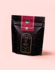 A black and gold resealable pouch labeled "Magic Hour" with a celestial design holds caffeine-free Ruby Moon™: Hibiscus Elderberry Tea, rich in vitamin C, set against a light pink background.