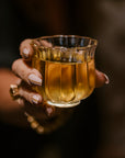 A hand with beige manicure and a gold ring holds an intricately designed, translucent glass filled with a golden liquid, possibly April 2024 Harvest - Grand Cru First Flush Goomtee Estate Darjeeling FTGFOP1. The background is dark, making the glass and exquisite tea from Magic Hour the focal point of the image.