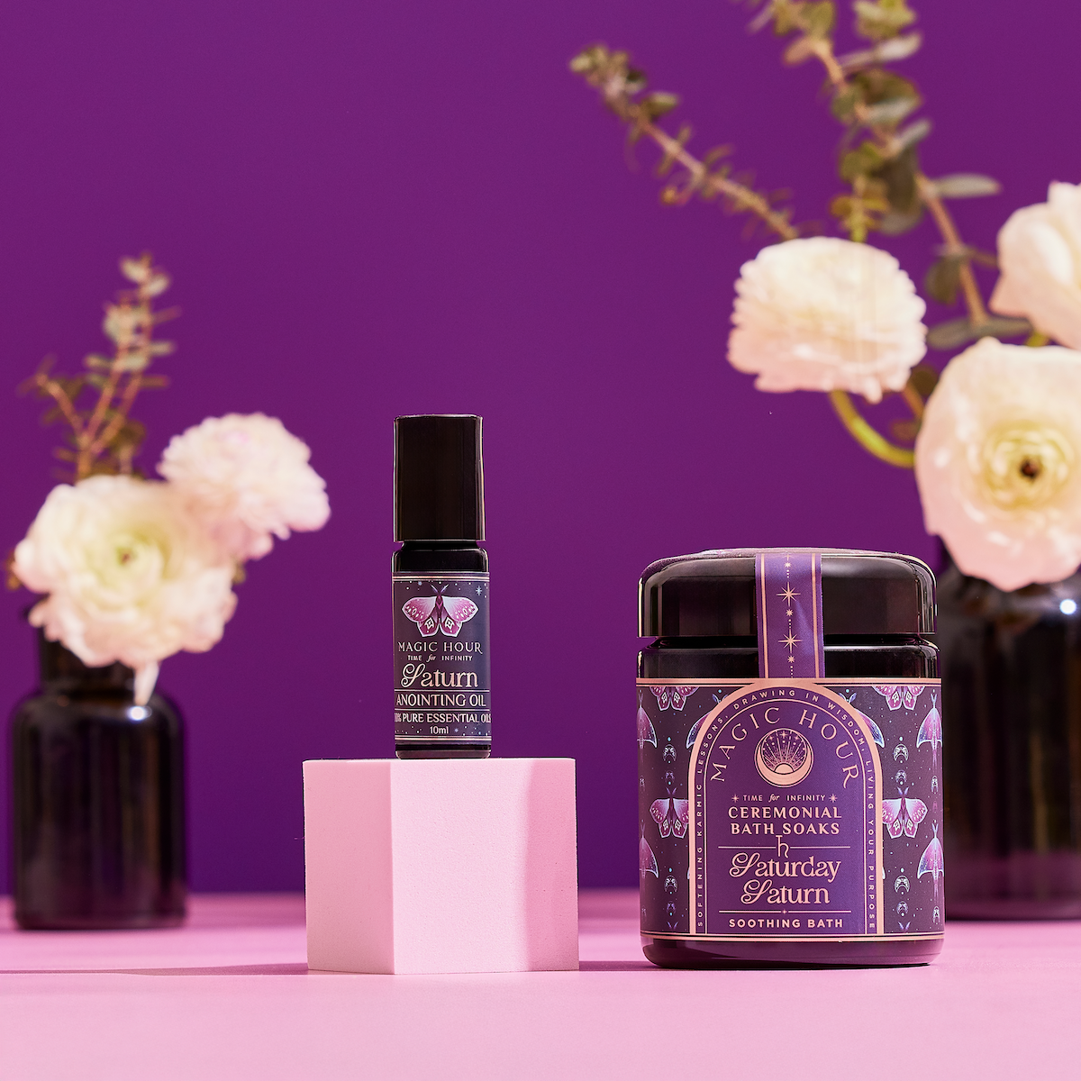 The Celestial Self-Care Ritual Set by Magic Hour, featuring a ceremonial bath soak and “Saturday Saturn” anointing oil, is displayed on a pink surface with white flowers—a celestial self-care ritual for your planetary bath.