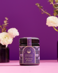 A jar of Ceremonial Bath Salt Soaks in a violet glass jar is centered on a pink surface with a purple backdrop, ideal for self-care rituals that inspire a celestial connection, flanked by blurred vases of white flowers.
