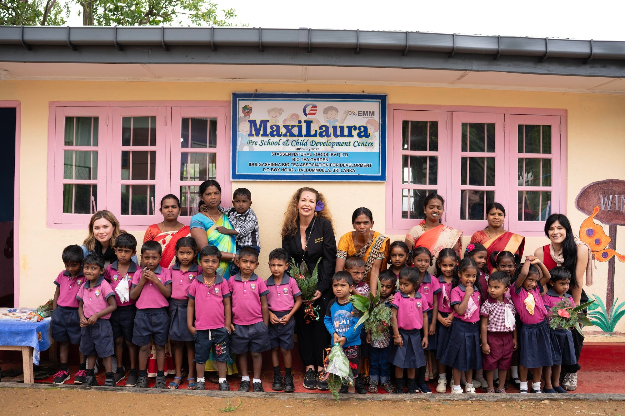 A group of children in matching school uniforms stand in front of a building labeled "MaxiLaura - The School of Child Development Centre." They are accompanied by several adults, including teachers and visitors, standing on either side of the group.
