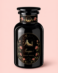 A black glass bottle labeled “Scorpio Tea for Sensual Brilliance” is adorned with gold floral motifs and an illustration of a figure beneath stars and a crescent moon, set against a light pink background—an enchanting homage to the mystical allure of Scorpio tea, delicately infused with notes evocative of rose congou and saffron black tea.