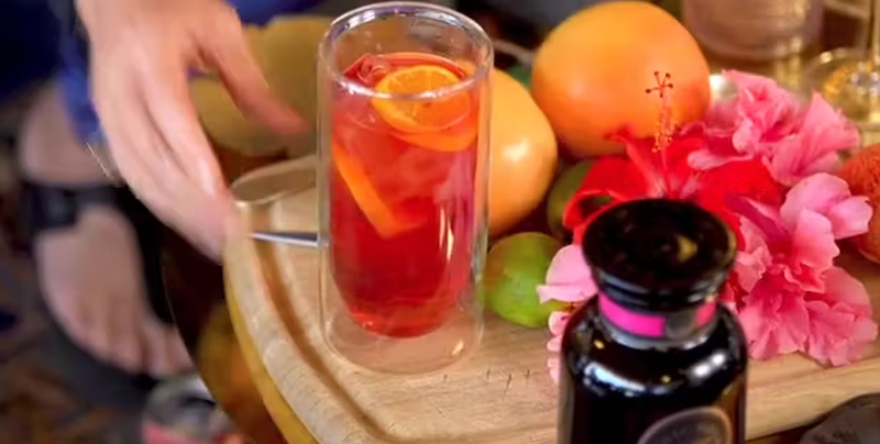 A hand reaches towards a tall glass filled with a bright red beverage, garnished with orange slices. The glass is on a wooden tray adorned with a variety of fruits including grapefruit, lime, and a pink hibiscus flower. A bottle with a black label is also on the tray.