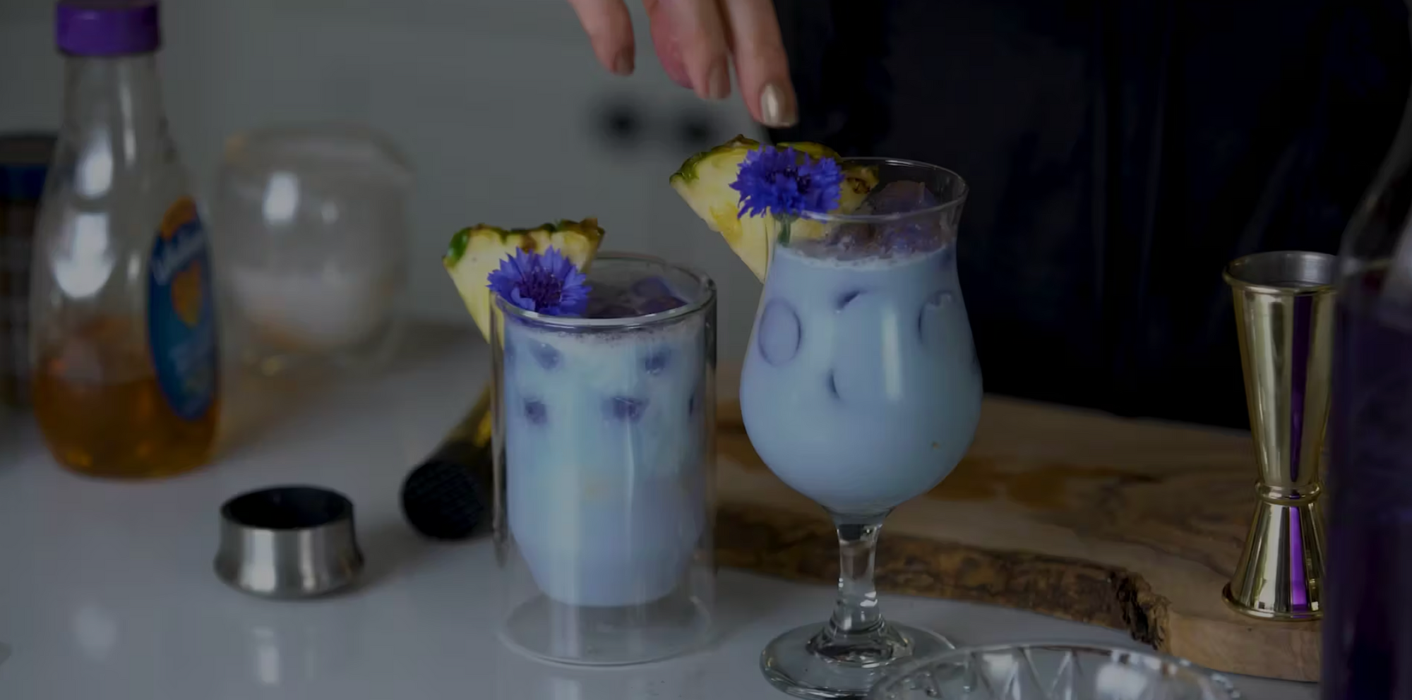 Two glasses of a blue-colored, milky beverage are topped with purple flowers and pineapple slices. A hand is gently placing a flower on one drink. Various bartending equipment and ingredients are seen on the white countertop in the background.