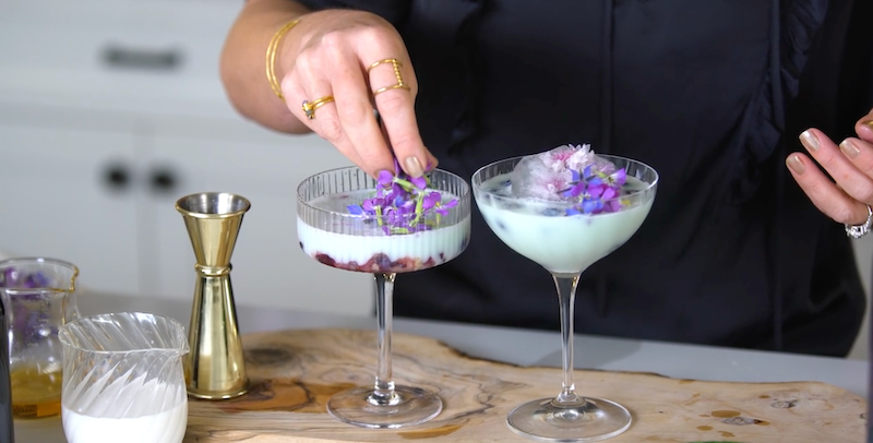 A person is delicately placing purple flowers onto two elegant cocktail glasses. The drinks are light-colored and garnished with clusters of violet petals. A gold cocktail measuring jigger, pitcher, and another container are on the wooden countertop.