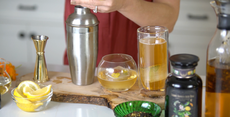 A person is using a cocktail shaker at a kitchen counter, with various ingredients and tools around. There are sliced lemons in a small bowl, a jigger, a green dish with dark contents, a jar with a label, and two glasses of a beverage, one tall and one round.