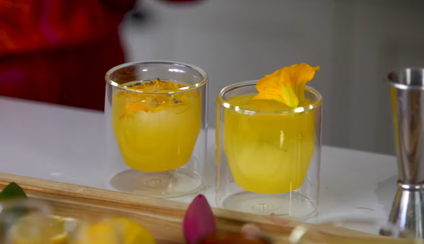 Two double-walled glass cups filled with a yellow-orange beverage are on display. One cup is garnished with small yellow flowers, and the other features a larger yellow flower. A metal jigger is visible on the right side of the image. Various ingredients surround.