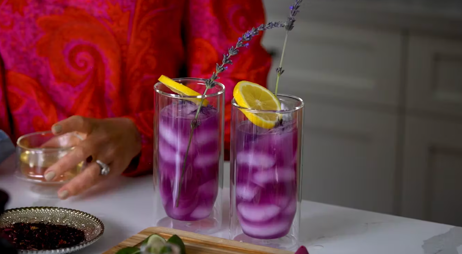 Two tall glasses filled with a purple beverage, ice cubes, a slice of lemon, and a sprig of lavender on the rim. A person in a red and pink patterned shirt is seen in the background, holding a small dish. The setting appears to be a kitchen.