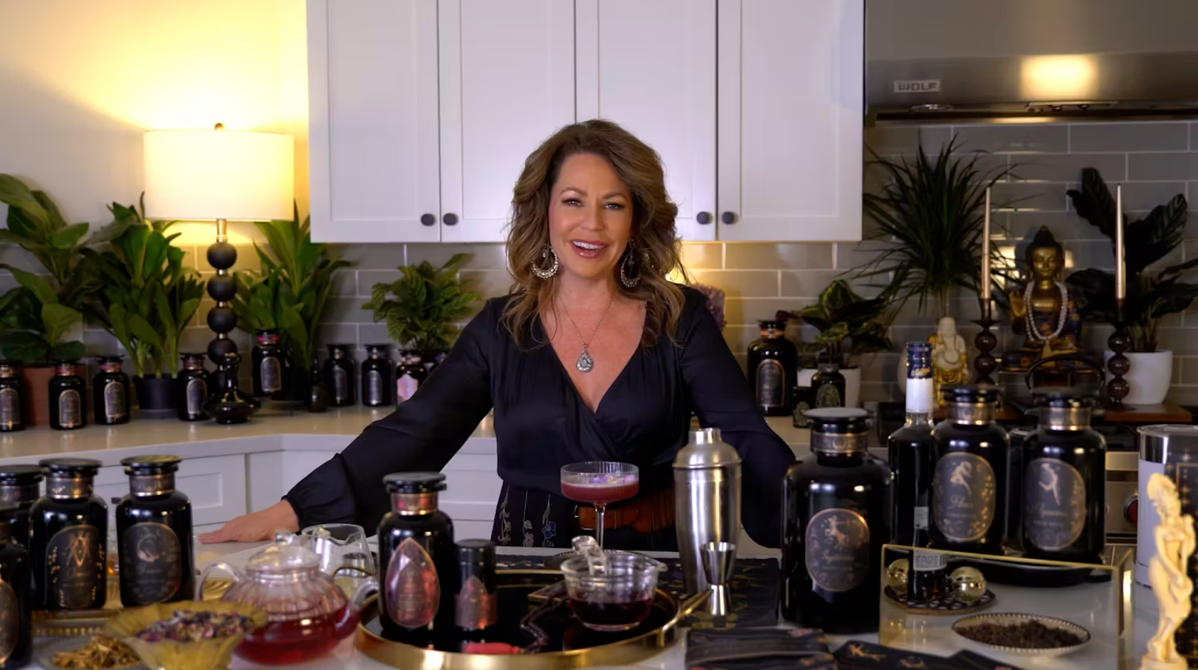 A woman stands confidently in a well-appointed kitchen, surrounded by an assortment of beautifully labeled bottles and ingredients. She wears a black top and smiles at the camera, with a prepared drink in front of her and various plants and decorative items in the background.