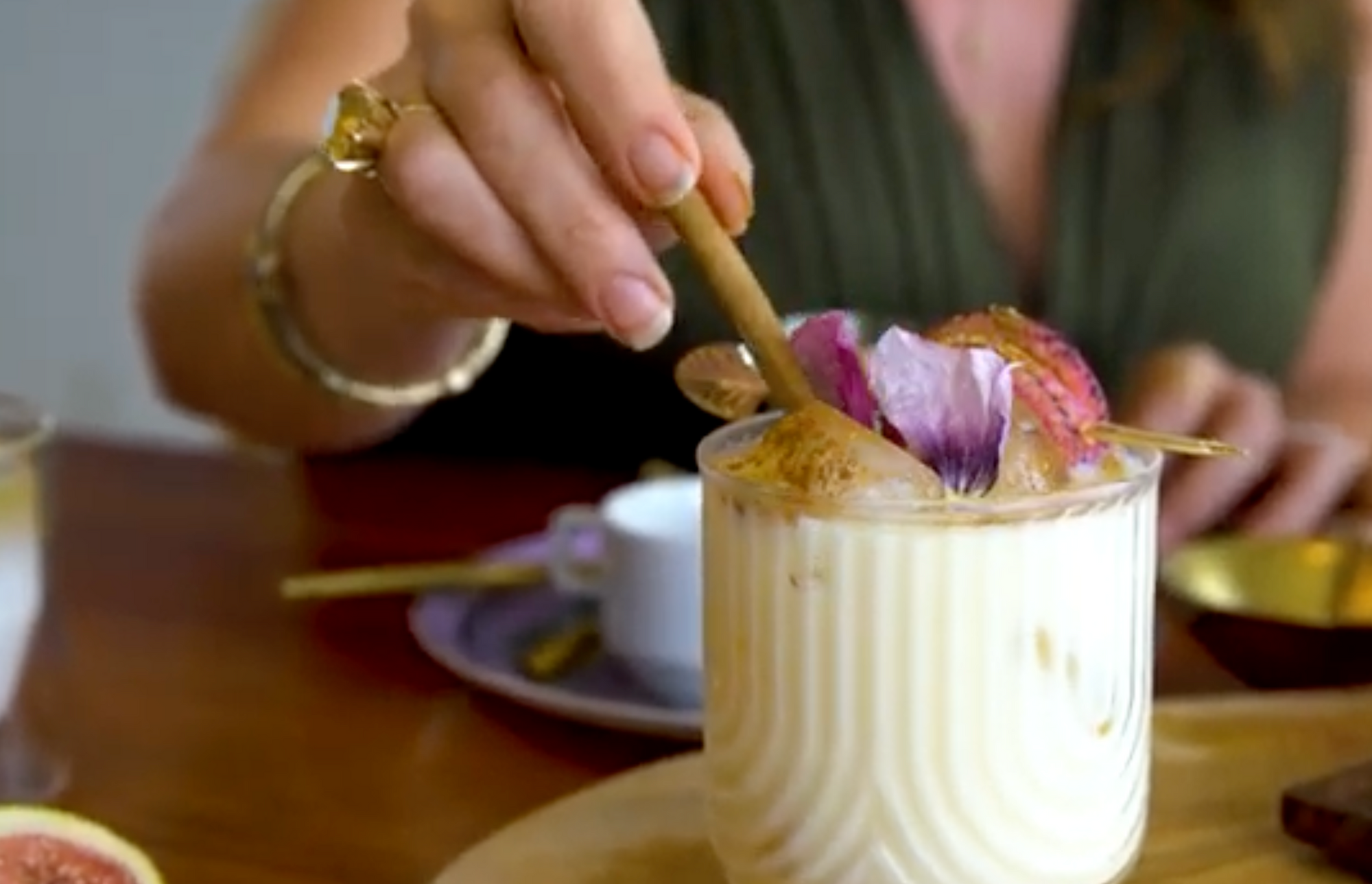 A person stirring a creamy drink topped with flower petals and cinnamon using a wooden spoon. The drink is in a glass placed on a wooden tray, and a fig is visible on the left side of the image. The person is wearing a bracelet and a ring.