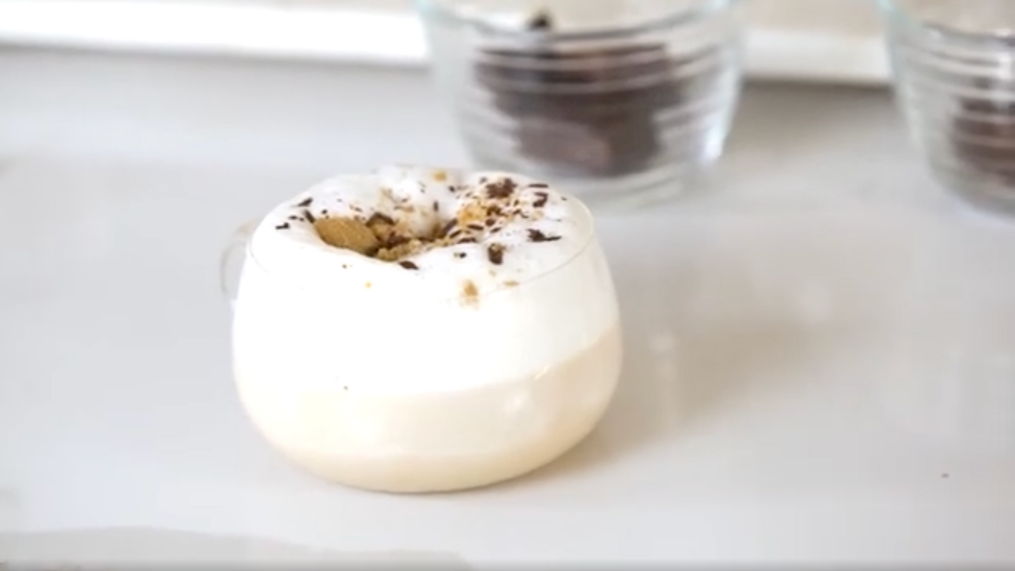 A small, clear glass cup filled with layered coffee is topped with thick foamy milk and sprinkled with chocolate or cocoa powder. The background has a white surface and some blurred out cups in the distance.