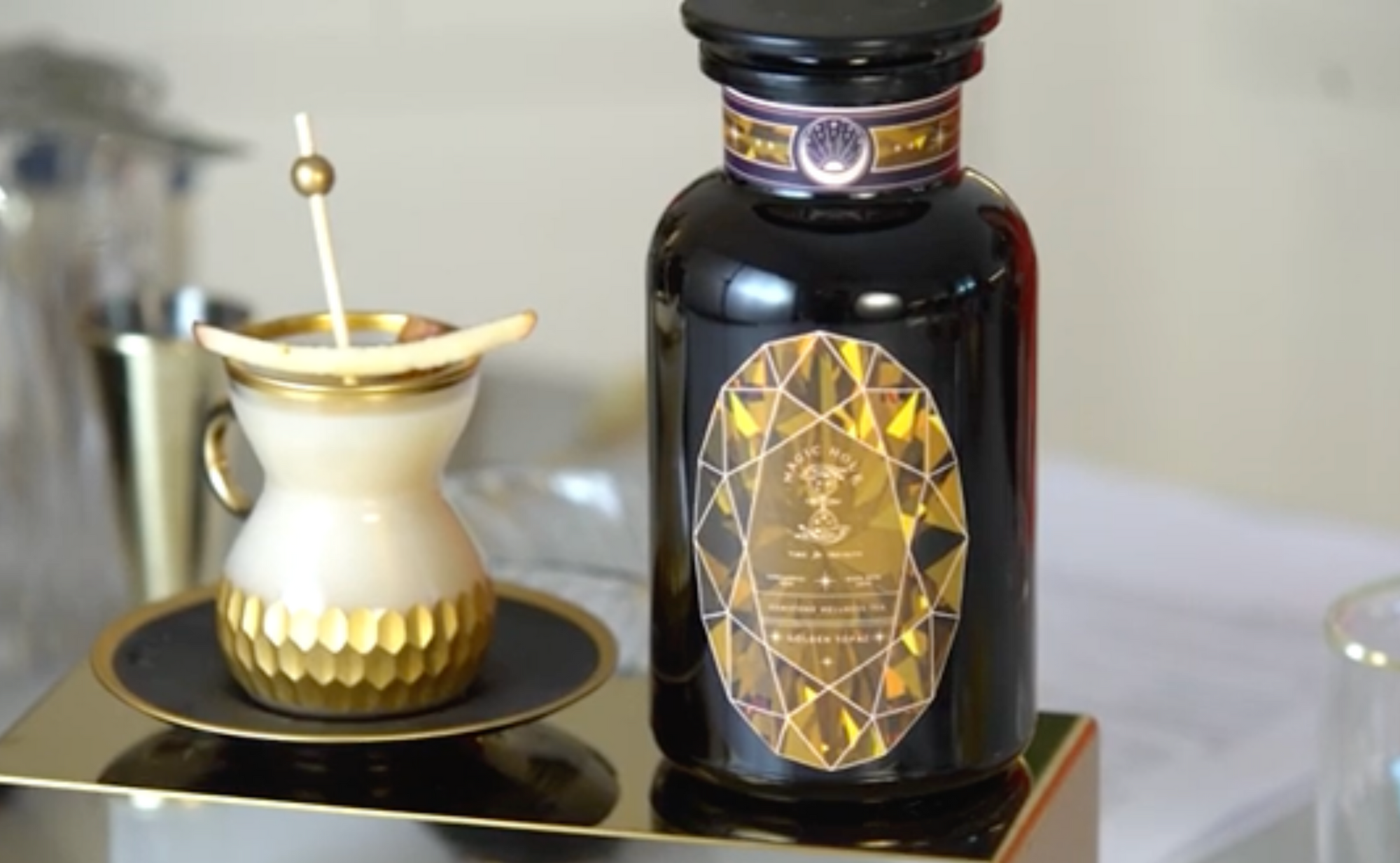 A luxurious black bottle with gold detailing and an intricate design label stands next to a cream and gold cup with a teaspoon sticking out. Both items are placed on a glossy black and gold platform, creating an elegant and refined display.