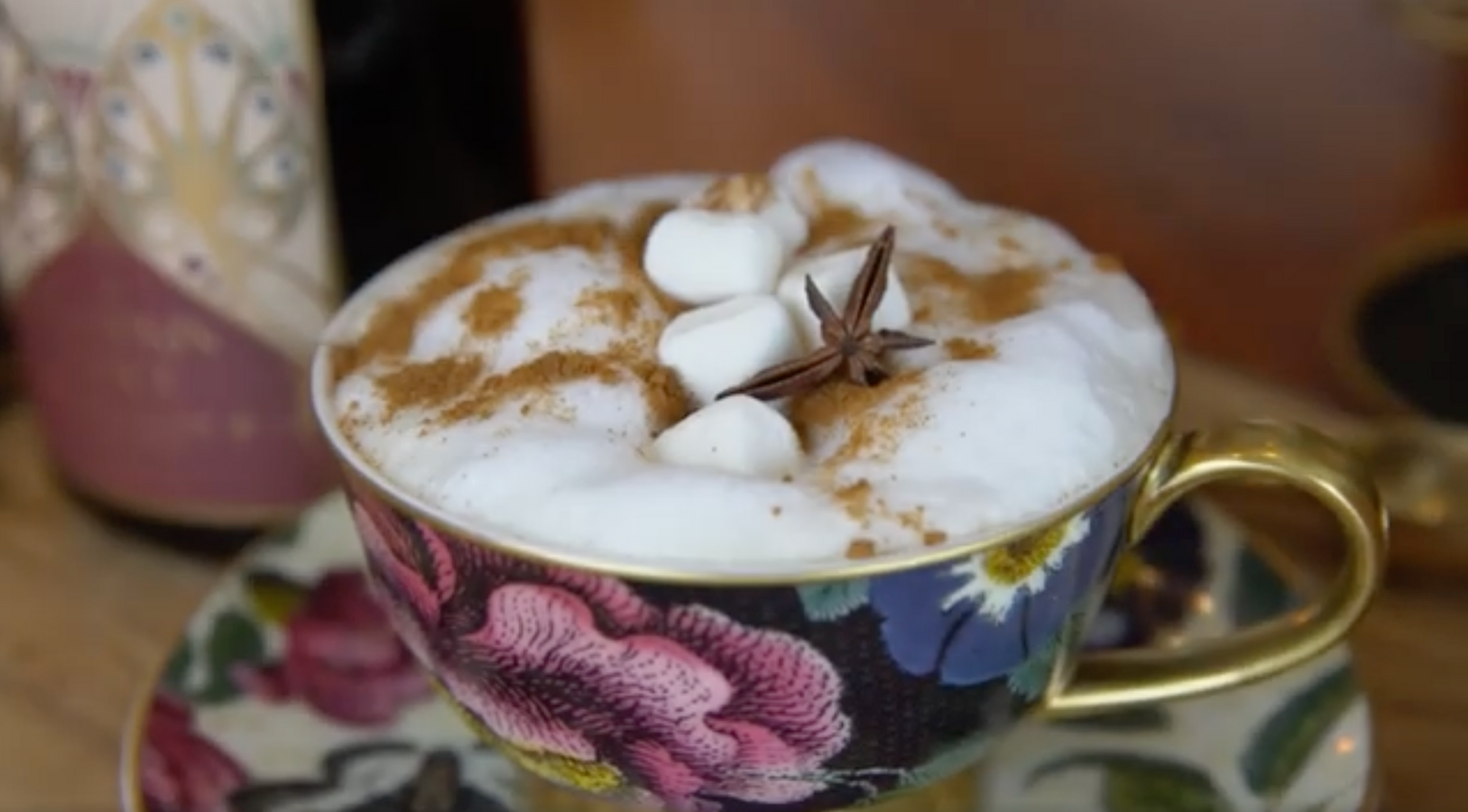 A floral teacup filled with frothy hot chocolate is topped with mini marshmallows, a sprinkle of cinnamon, and a star anise. The matching saucer is also visible, and there's a blurred background with a bottle and a wooden surface.