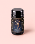 A black jar labeled “MAGIC HOUR” with an intricate design depicts a woman holding a wolf and a sword. The label reads “She Rises: Skadi The Sovereign: Winter Berry Puerh.” This striking berry-infused tea embodies the spirit of winter resilience. The jar is set against a light pink background.