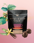 A black pouch labeled "Small Batch Magic Valentine’s 2026" stands upright amid hazelnuts, a vanilla flower, vanilla beans, and chocolate truffles—evoking the indulgent aroma of Madagascar vanilla rooibos—on a soft pink gradient background.