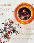 A glass cup of tea with raspberry, chocolate, and rose petals on a white surface. Loose leaf tea leaves and rose petals are scattered next to the cup. Text on the image reads: "Soulmate: Chocolate-Raspberry-Rose Black Tea Traveler" and "Energizes, Aids Digestion, Supports Heart Health." Savor the Magic Hour tea experience.