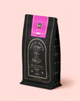 A black and gold tea pouch labeled "Soulmate: Chocolate-Raspberry-Rose Black Tea Refill" with an hourglass illustration and pink seal stands upright on a light pink background, suggesting the enchanting organic blend inside.