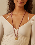 A woman with curly hair, wearing a cream-colored off-shoulder top, is shown from the shoulders up. She wears a Magic Hour Spiritual Evolution Rhodonite Mala with a circular, intricate pendant hanging at the end. She stands against a beige background.