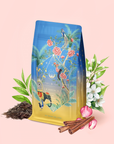 A vibrant Sri Lankan Sunrise tea package adorned with floral and bird illustrations stands amid tea leaves, cinnamon sticks, white blossoms, and pink petals on a pale pink background—perfectly capturing the essence of our organic black tea cardamom cinnamon blend.