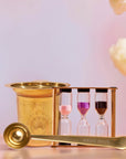 Loose Leaf Tea Starter Set: Includes a gold tea strainer, matching spoon, and a three-hourglass tea timer with pink, purple, and brown sands—all beautifully arranged on a light surface with blurred flowers in the background. This elegant set is perfect for brewing your favorite loose-leaf tea.