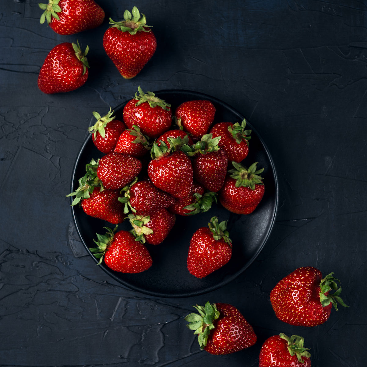 A black plate filled with fresh, red strawberries is placed on a dark textured surface. Several strawberries are scattered around the plate, creating a vibrant contrast with the background.