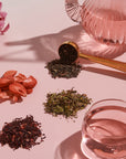 A pink tea set up with loose Strawberry Shortcake Iced Tea with Hibiscus leaves from Magic Hour and dried strawberries on display. A glass teapot and teacup filled with pink liquid are complemented by a gold tea scoop. Pink flowers decorate the background, adding an elegant touch to the arrangement.