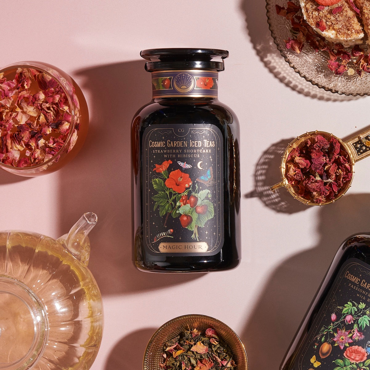 A bottle labeled "Magic Hour Strawberry Shortcake Iced Tea with Hibiscus" sits on a pink surface surrounded by various dishes containing tea leaves, dried flowers, and Organic Freeze Dried Strawberries. The dark bottle features colorful floral artwork. Other tea containers and a teapot are partially visible. Iced Tea.