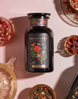 A bottle labeled "Magic Hour Strawberry Shortcake Iced Tea with Hibiscus" sits on a pink surface surrounded by various dishes containing tea leaves, dried flowers, and Organic Freeze Dried Strawberries. The dark bottle features colorful floral artwork. Other tea containers and a teapot are partially visible. Iced Tea.