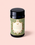 A jar of Flower Powder: Strawberry Matcha, featuring a green and floral label, rests on a light pink background, showcasing this beauty food blend crafted from premium ceremonial matcha and juicy strawberries.