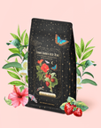 A tea bag labeled "Strawberry Shortcake Iced Tea with Hibiscus" features cosmic designs and strawberries, surrounded by illustrated flowers, leaves, and strawberries on a light pink background. The packaging includes a butterfly, capturing the delightful essence of creamy strawberry shortcake in every detail.