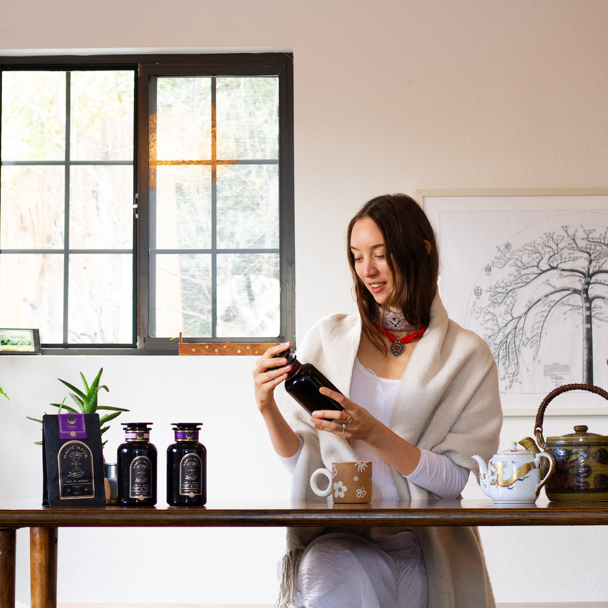 A woman in a white outfit and shawl sits at a wooden table, examining a dark bottle. The table holds other dark jars, a tea set, and a potted plant. A window and a framed tree illustration are visible in the background.