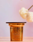 The Steep The Stars - Artisan Tea Strainer, featuring elegant engraved patterns, rests on a light pink surface with blurred white flowers and a soft pink backdrop, perfect for enjoying loose leaf tea.