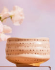 The Ceremonial Matcha Bowl, a handmade cream ceramic with star-shaped cutouts, rests on glass beside pink flowers against a pastel pink backdrop.