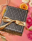 The Summer Solstice Mystery Box—a black gift box with gold stars and a "MAGIC HOUR" gold ribbon—is accented by pink flowers, an orange slice, and pearls. It includes exclusive, limited-edition tea samplers perfect for a magical summer surprise.