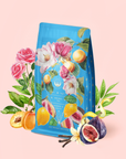A blue "Summer in Amalfi" tea package adorned with illustrated flowers, peaches, figs, and leaves, surrounded by matching fruits and blossoms on a soft pink background. The design evokes the refreshing blend inside—green tea oolong enhanced with organic lemon peel.