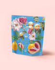 A blue resealable pouch of "Summer in Amalfi" stands upright against a pale pink background, featuring colorful illustrations of flowers, lemons, figs, and cashew fruits that highlight its Mediterranean Tea blend with Lemon Bergamot.