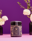 A Violet Glass Jar of Ceremonial Bath Salt Soaks is showcased on a pink surface, with dark vases of white flowers and greenery against a vibrant purple background, creating a celestial vibe.