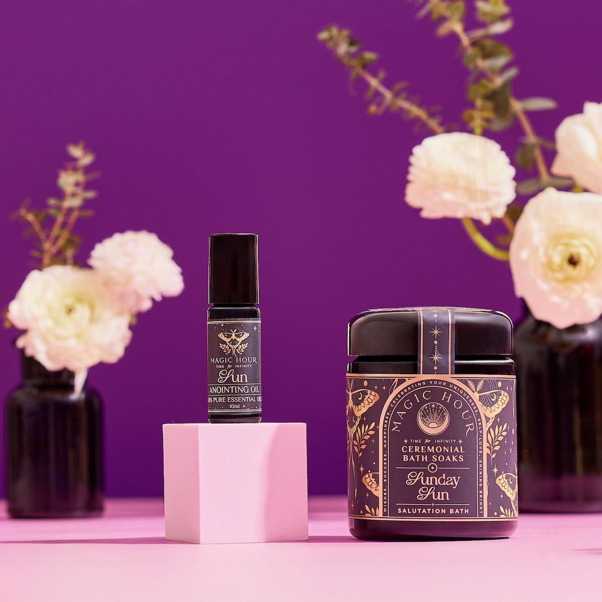 The Celestial Self-Care Ritual Set is displayed on a pink surface with a purple background. Cream-colored flowers in dark vases blur behind, highlighting the ornate black and gold labels—perfect for celestial self-care.