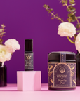 The Celestial Self-Care Ritual Set is displayed on a pink surface with a purple background. Cream-colored flowers in dark vases blur behind, highlighting the ornate black and gold labels—perfect for celestial self-care.