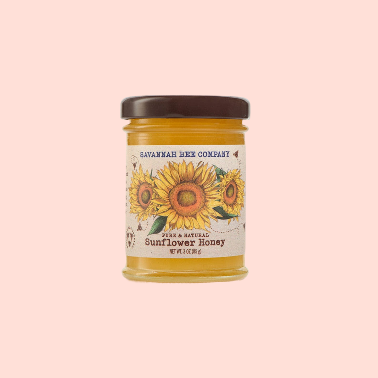 A jar of Savannah Bee Co. Honey - Sunflower, labeled "Pure & Natural Sunflower Honey, Net Wt. 3 oz (85 g)" with sunflower imagery, sits against a pink background. This golden delight is crafted by dedicated beekeepers inspired by Ukraine's sunflower fields.