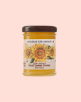 A jar of Savannah Bee Co. Honey - Sunflower, labeled "Pure & Natural Sunflower Honey, Net Wt. 3 oz (85 g)" with sunflower imagery, sits against a pink background. This golden delight is crafted by dedicated beekeepers inspired by Ukraine's sunflower fields.